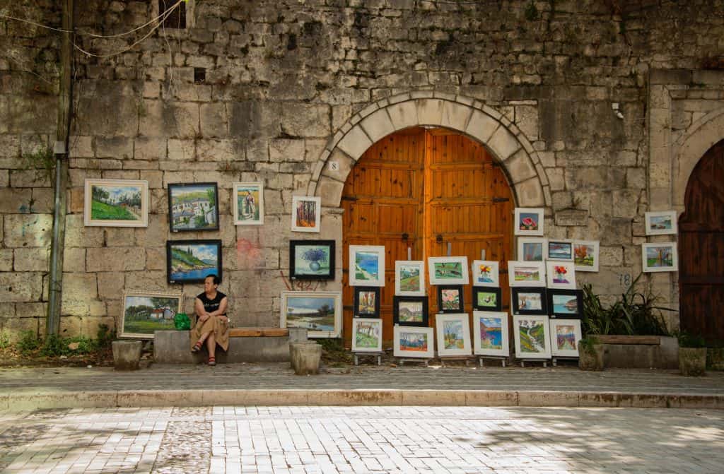 Art and frames being sold on the street against an old wall in Tirana, with a seated person looking away from the camera