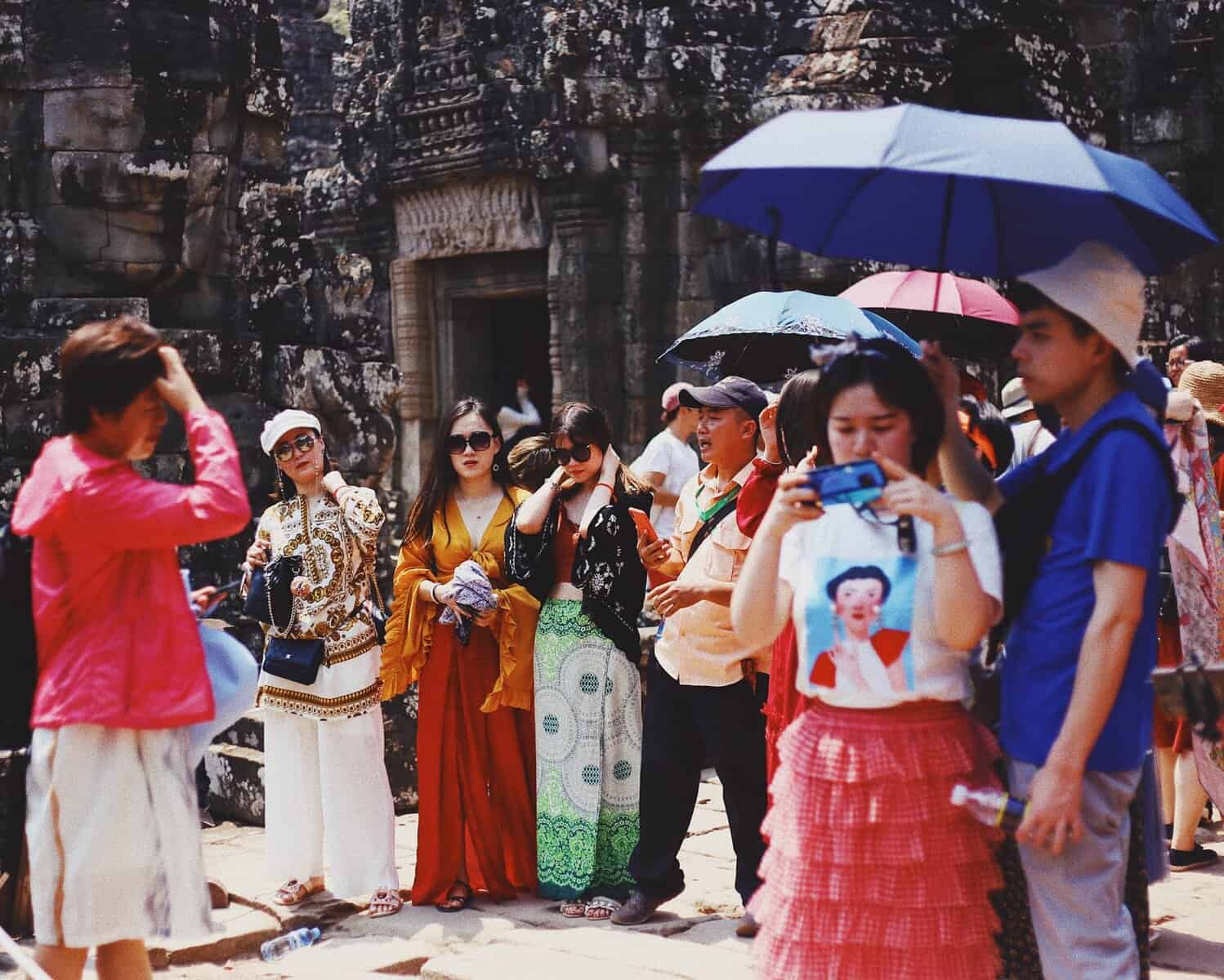 Group of tourists standing outside a temple in Cambodia. Some are adjusting their clothes, some are taking photos, while some are standing under the shade of umbrellas.