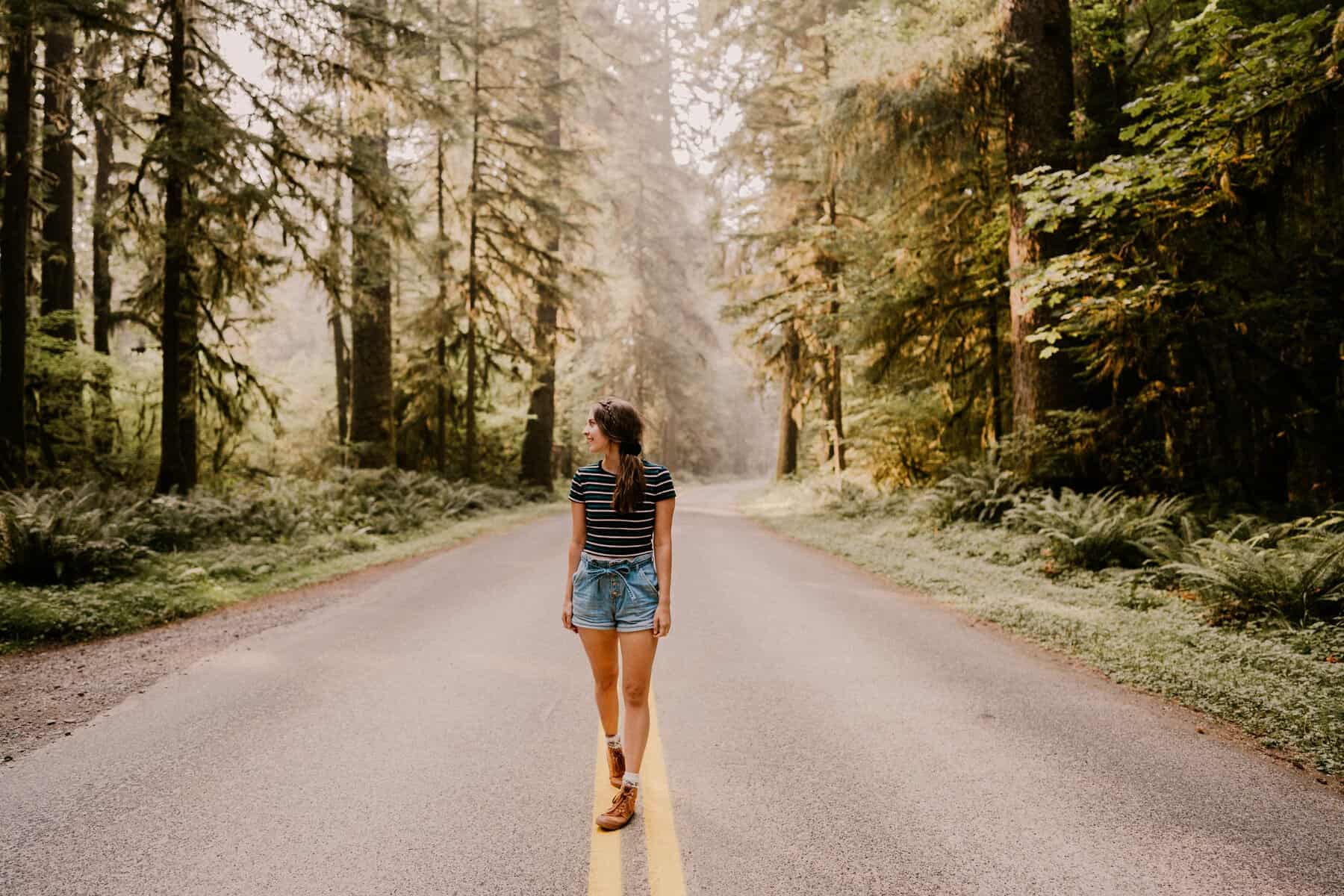 Woman in black and white striped shirt and denim shorts standing in the middle of the road with trees on both sides, smiling