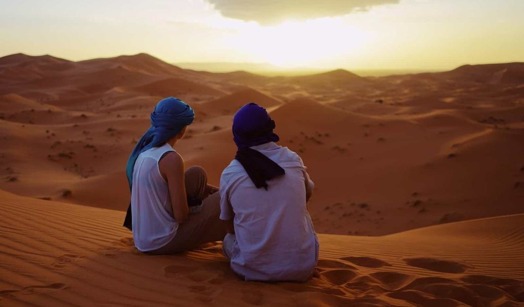 Man and woman with head gear looking at a sunset over a desert in Morocco.